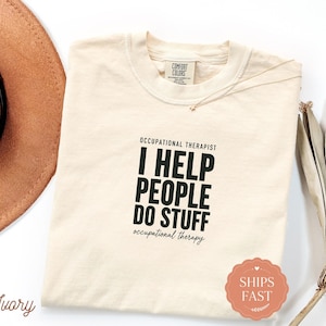 May include: An ivory-coloured short-sleeve t-shirt featuring black text that reads OCCUPATIONAL THERAPIST I HELP PEOPLE DO STUFF occupational therapy. The shirt has a crew neckline and a Comfort Colors brand label printed on the inside collar.