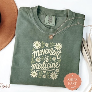May include: A moss green short-sleeve t-shirt featuring a graphic print of white daisies and green leaves surrounding the text "movement is medicine" in a cursive font. The shirt has a crew neckline and a Comfort Colors brand label at the neck.
