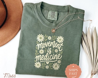 May include: A moss green short-sleeve t-shirt featuring a graphic print of white daisies and green leaves surrounding the text "movement is medicine" in a cursive font. The shirt has a crew neckline and a Comfort Colors brand label at the neck.