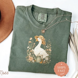 May include: A moss green short-sleeved t-shirt featuring a graphic of a white goose wearing a straw hat and holding a small watering can, surrounded by various flowers. The shirt has a Comfort Colors label at the neck and a crew neckline.