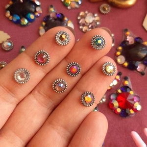 May include: A hand displaying a variety of small, round, iridescent rhinestones with silver settings. The rhinestones are arranged in a row on the fingers, with each stone having a different colour.
