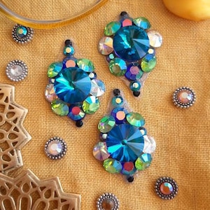 May include: Three decorative pendants with large, faceted blue gemstones and iridescent embellishments. Small, decorative studs and a yellow rubber duck are scattered on a yellow fabric background.