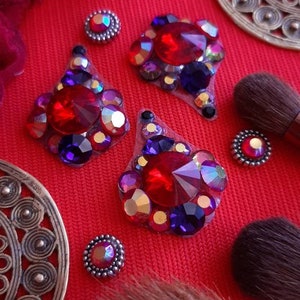 May include: Assortment of red, purple and gold rhinestone pasties with a silver setting. The pasties are on a red surface with makeup brushes and a gold metal earring.
