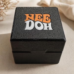 Könnte beinhalten: Eine schwarze Box mit dem Text "NEE DOH" in orangefarbenen und weißen Buchstaben. Die Box hat eine strukturierte Oberfläche und einen Klappdeckel. Der Hintergrund ist neutral.