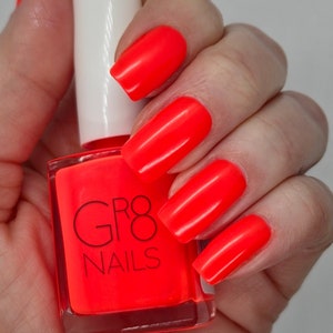 May include: A hand with long, bright neon orange nails next to a bottle of matching nail polish. The nail polish bottle is square with a white cap and the words "GR8 NAILS" in black.