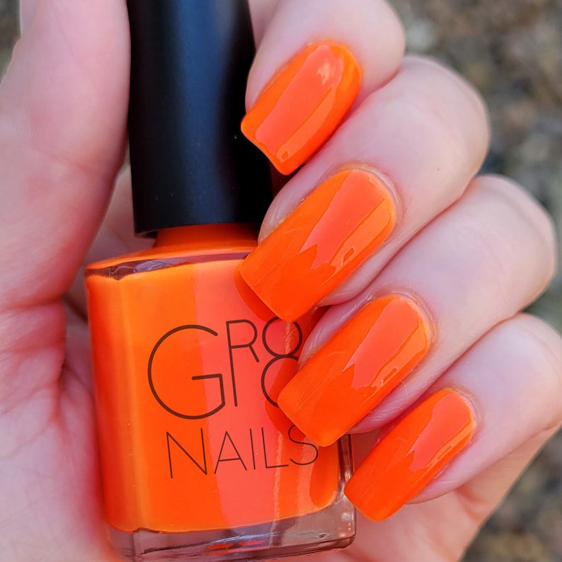 May include: A bottle of bright orange nail polish with a black cap is held in a hand with matching orange polished nails. The nail polish bottle has the brand name "GRO8 NAILS" printed on it.