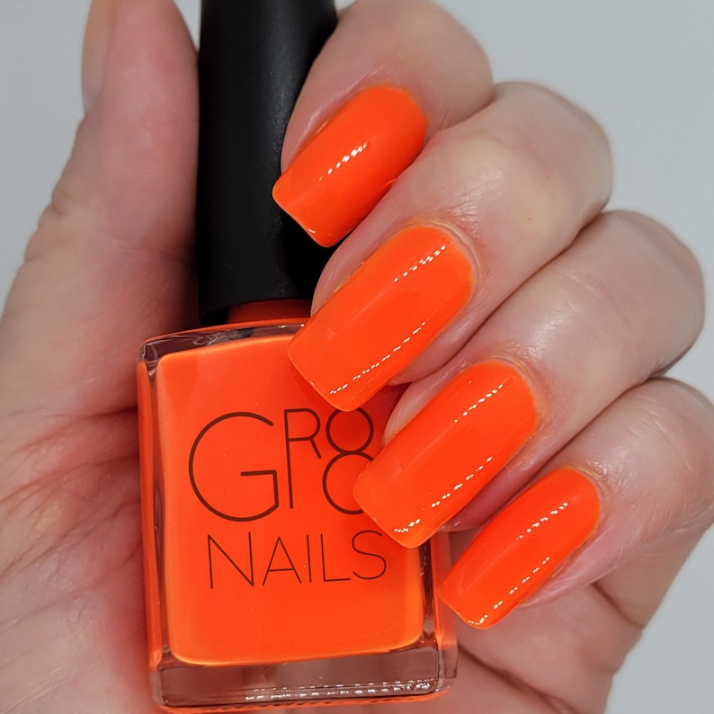 May include: A bottle of bright orange nail polish with a black cap is held in a hand. The nails are painted the same vibrant orange color. The bottle has the words "GRO8 NAILS" printed on it.