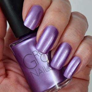 May include: Close-up of a hand with polished nails in a metallic lavender shade. The nail polish bottle is also lavender, with the brand name "GRO NAILS" visible. The nails have a glossy finish.