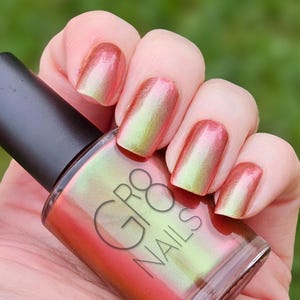 May include: A bottle of GR8 NAILS nail polish with a black cap is held in a hand. The nails are painted with a color-shifting polish that transitions from a reddish-orange to a light green. The background is a blurred green.
