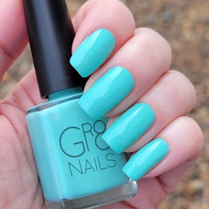 May include: A bottle of turquoise nail polish with a black cap, held by a hand with matching turquoise painted nails. The bottle has the brand name "GRO NAILS" printed on it.