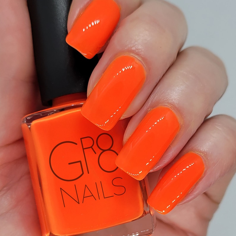 May include: Bright orange nail polish and painted nails. The square bottle of nail polish has a black cap and the words "GR8 NAILS" printed on the front. The nails are a vibrant, glossy orange.