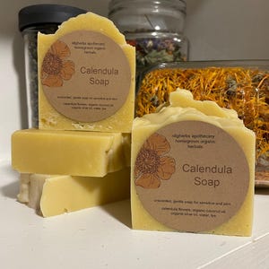 May include: Two bars of yellow calendula soap with brown labels that read "idigherbs apothecary homegrown organic herbals Calendula Soap" and list the ingredients. The soap is sitting on a white surface.