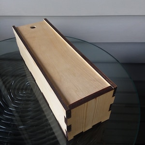 Puede incluir: Una caja de madera con un borde marrón oscuro y un pequeño agujero en la parte superior. La caja está hecha de madera clara y tiene un acabado liso.