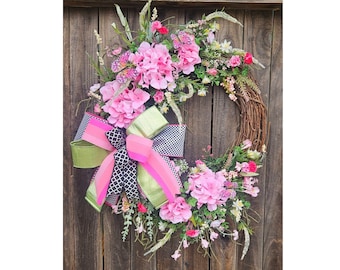 May include: A pink and green floral wreath with a black and white checkered bow. The wreath is made of grapevine and is decorated with pink hydrangeas, pink and white flowers, and green leaves.