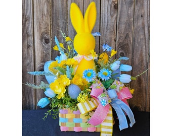 May include: A yellow bunny sits in a pastel colored basket filled with blue and yellow flowers. The basket is decorated with a pink and blue bow.