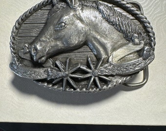 May include: Silver-toned belt buckle featuring a horse's head with a rope design around the mane and a stylized spur design below.
