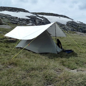 May include: A white tarp tent pitched in a grassy field with a rocky mountain in the background. The tent is set up with a single pole and guy lines.