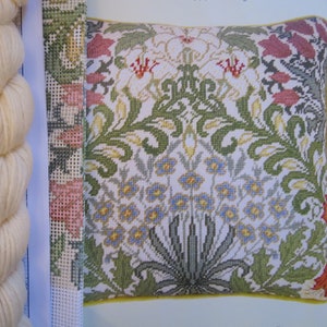 Peut inclure: Un gros plan d'un coussin en point d'aiguille avec un motif floral. Le coussin est blanc avec des fleurs vertes, jaunes, bleues et roses. Le coussin est entouré d'une bordure jaune.