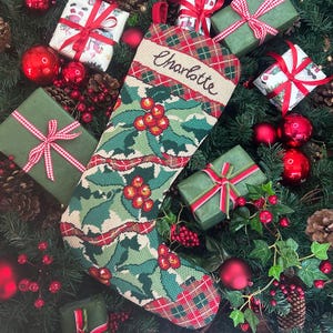 Könnte beinhalten: Ein Weihnachtsstrumpf mit einem grünen Stechpalmen- und roten Beerenmuster, mit dem Namen "Charlotte" auf der Oberseite gestickt. Der Strumpf ist von verpackten Geschenken, roten Kugeln und Tannenzapfen umgeben und schafft eine festliche Urlaubsszene.