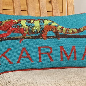 May include: Teal rectangular pillow with a red border. The pillow features a colorful chameleon perched on a branch, with the word "KARMA" in red block letters below the chameleon. The chameleon has red, yellow, and green markings.