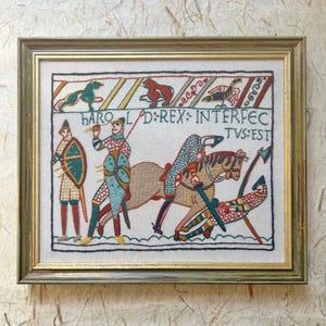 May include: A framed embroidery depicting a scene from the Bayeux Tapestry with figures in chainmail, horses, and the text HAROLD REX INTERFECTUS EST. The artwork features red, blue, green, and tan thread on a light fabric, enclosed in a gold frame.