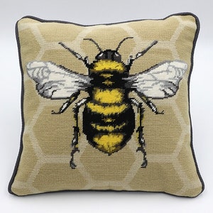 May include: A beige cushion with a black border features an embroidered image of a bee with yellow and black stripes and white wings. The bee is centred on the cushion and is surrounded by a honeycomb pattern.