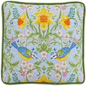 Könnte beinhalten: Quadratisches Zierkissen mit hellblauem Hintergrund mit einem Blumen- und Vogeldesign. Das Design umfasst gelbe Narzissen, rosa Blüten und blaue Vögel. Das Kissen hat einen grünen Rand.