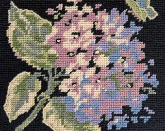 以下が含まれることがあります： 紫と青の花が緑の葉と蝶で囲まれた、針仕事のデザインのクローズアップ。蝶は右上の隅にあります。