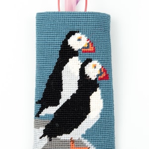 May include: A blue needlepoint glasses case featuring a design of two puffins on a white background. The puffins are black, white and orange. A pair of pink glasses is sticking out of the top of the case.