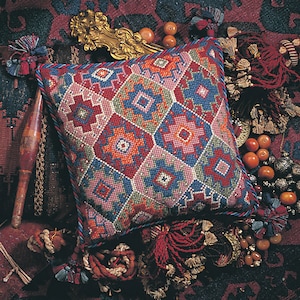 Puede incluir: Una almohada cuadrada colorida con un patrón geométrico en tonos rojos, azules, verdes y rosas. La almohada está rodeada de borlas y cuentas.