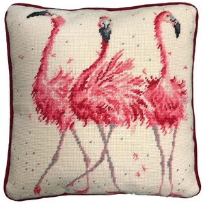 May include: A white pillow with a red border features a needlepoint design of three pink flamingos standing on a white background with scattered pink dots.
