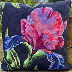May include: Navy blue cushion with a vibrant floral design. The flower is depicted in pink, red, purple, and blue hues, with green leaves and stems. The cushion is square and appears to be made from a woven material.