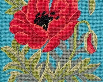 以下が含まれることがあります： 青い背景に赤いポピーの花が緑の葉と茎で描かれています。花は赤、緑、灰色の糸で刺繍されています。