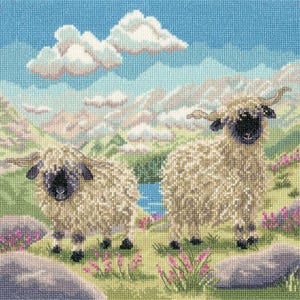 May include: Cross-stitch image of two sheep with curly, light-coloured wool and black faces, set in a green field with pink flowers. The background depicts mountains, a blue river, and a cloudy sky.