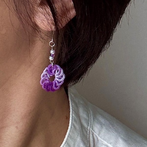 May include: A close-up of a single earring. The earring features a circular, woven design in shades of purple and white, with a silver hook and small, clear beads. The earring is worn on the ear.