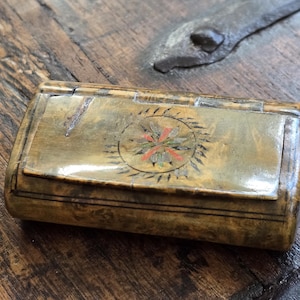 May include: A rectangular, tan-coloured box with a hinged lid featuring a central circular motif with red and green floral-like details. The surface has a mottled, wood-grain appearance with dark brown linear accents along the edges and a smooth finish.