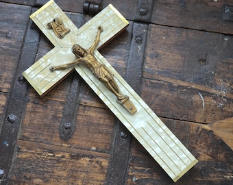 Peut inclure: Une croix ancienne avec une figurine de Jésus dorée sur une croix. La croix présente une incrustation crème à motifs avec une bordure dorée. La croix est posée sur un fond en bois sombre.