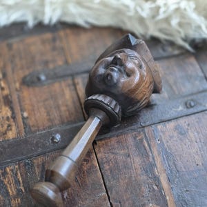 Puede incluir: Un tapón de botella de madera tallado a mano, de época, que presenta un rostro detallado con un sombrero. El tapón es de color marrón oscuro y tiene un mango largo y delgado. El fondo es una superficie de madera rústica.