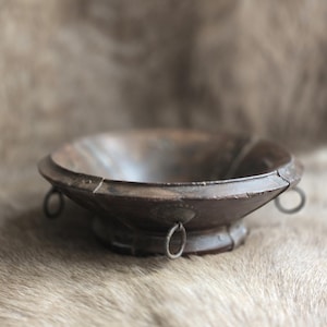 May include: A dark brown, antique-style wooden bowl with a wide, shallow design. The bowl features a circular base and a flared rim, with small metal ring handles attached to the sides. The bowl is set against a textured, neutral-toned background.
