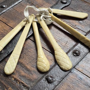 Könnte beinhalten: Eine Sammlung antiker, elfenbeinfarbener Werkzeuge, möglicherweise für den Küchengebrauch, die mit einer weißen Schnur zusammengebunden sind. Die Werkzeuge weisen geschnitzte Details auf, darunter Rauten- und Rippenmuster. Sie liegen auf einer dunklen Holzoberfläche mit Metallakzenten.
