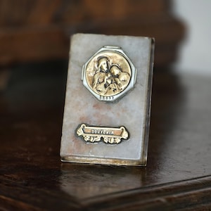 May include: A small, rectangular religious souvenir with a marble-like finish. It features a gold and silver emblem depicting a religious scene and a banner that reads "SOUVENIR". The item is resting on a dark wooden surface.