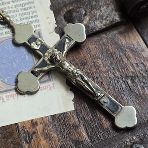 May include: A detailed, antique crucifix with a silver-toned Christ figure and a dark blue cross. The cross has decorative, rounded ends and is set against a backdrop of aged paper and wood.