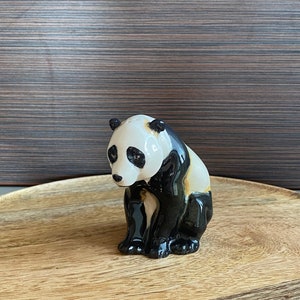 May include: A ceramic panda figurine in black and white. The panda is depicted in a seated position, with its head gently lowered. The figurine is placed on a wooden tray.