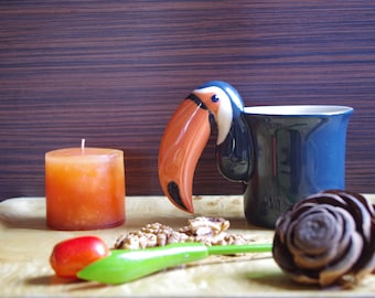 Puede incluir: Naturaleza muerta con una taza azul con asa de pico de tucán, una vela naranja, nueces, una fruta roja, un objeto verde y una piña decorativa. Los objetos están dispuestos sobre una bandeja de madera.