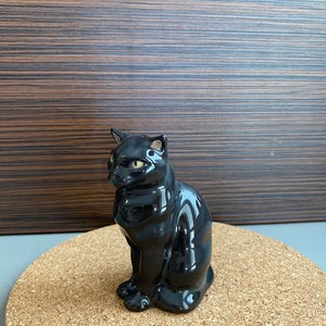 Puede incluir: Figura de gato de cerámica negra brillante con ojos amarillos, sentado. El gato tiene un acabado pulido y está colocado sobre una superficie redonda de corcho. El fondo es un panel de madera.