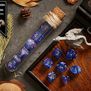 May include: A set of blue and pink iridescent dice with gold numbers. The dice are in a glass test tube with a cork stopper and twine. The dice are on a wooden surface with a small dragon figurine.