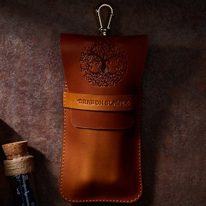 May include: A brown leather case with a tree of life design and the text "Dragon Slayer". The case has a gold metal hook.