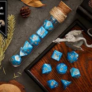 May include: A set of blue and gold dice with a sky marble finish. The dice are in a glass tube with a cork stopper and are on a wooden surface. The dice are labeled with numbers 1 through 20.