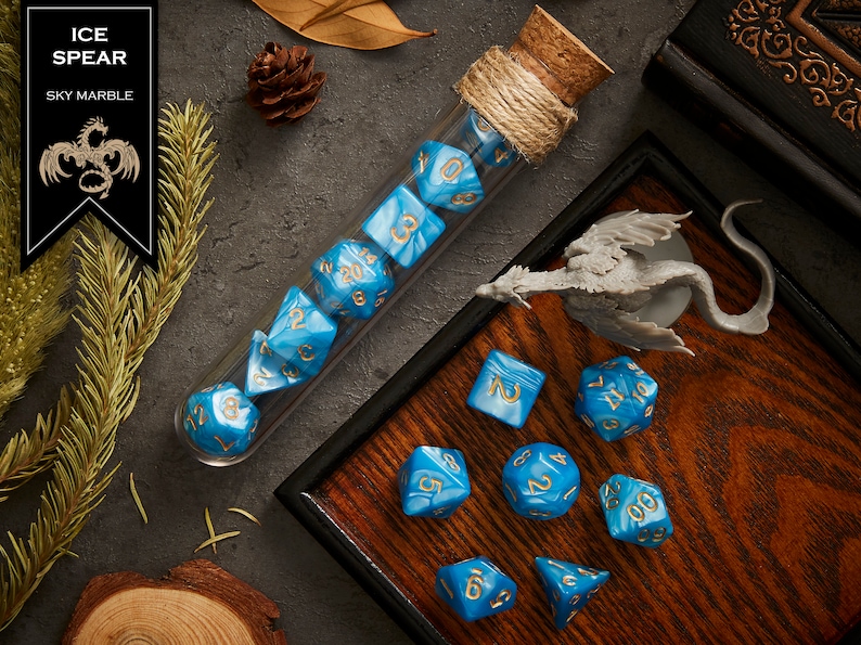 May include: A set of blue and gold dice with a sky marble finish. The dice are in a glass tube with a cork stopper and are on a wooden surface. The dice are labeled with numbers 1 through 20.