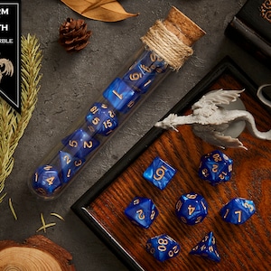 May include: A set of blue and gold dice with a dragon logo, "Storm of Death", and "Blue Marble" text. The dice are in a glass test tube with a cork stopper and are on a wooden surface with a miniature dragon figurine.